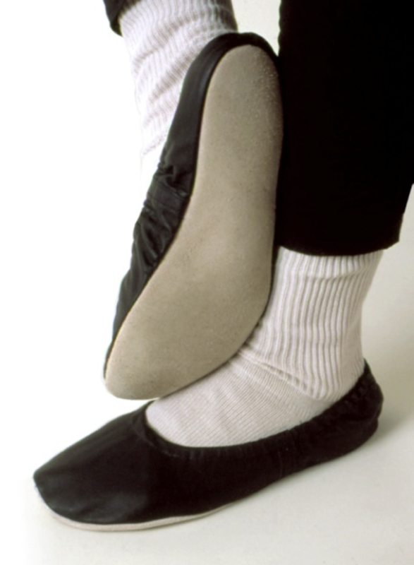 Close-up of a dancer's feet positioned in third position wearing black ballet slippers and white, knitted leg warmers. The dancer's right foot is flat on the ground while the left foot is on demi-pointe, showing the suede sole of the slipper.